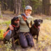 A woman kneeling in a field with her arms around her two black labs